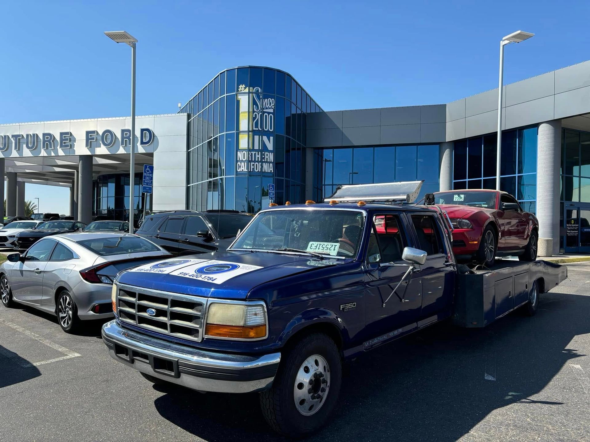 Blue flatbed tow truck parked outside a car dealership on a sunny day.