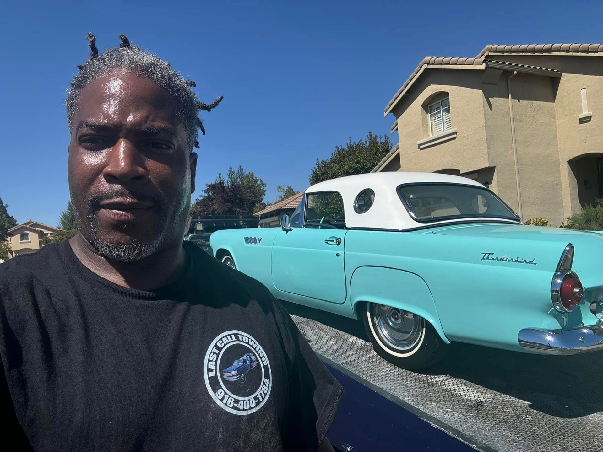 Man taking a selfie beside a turquoise classic car parked on a driveway in front of a beige house