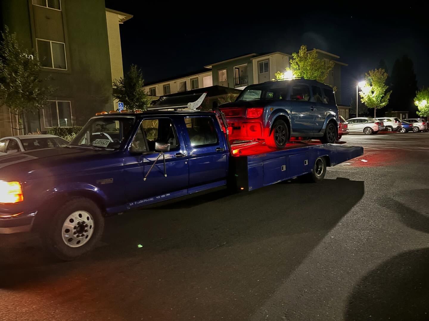 Blue tow truck hauling a red SUV on a flatbed at night in a parking lot