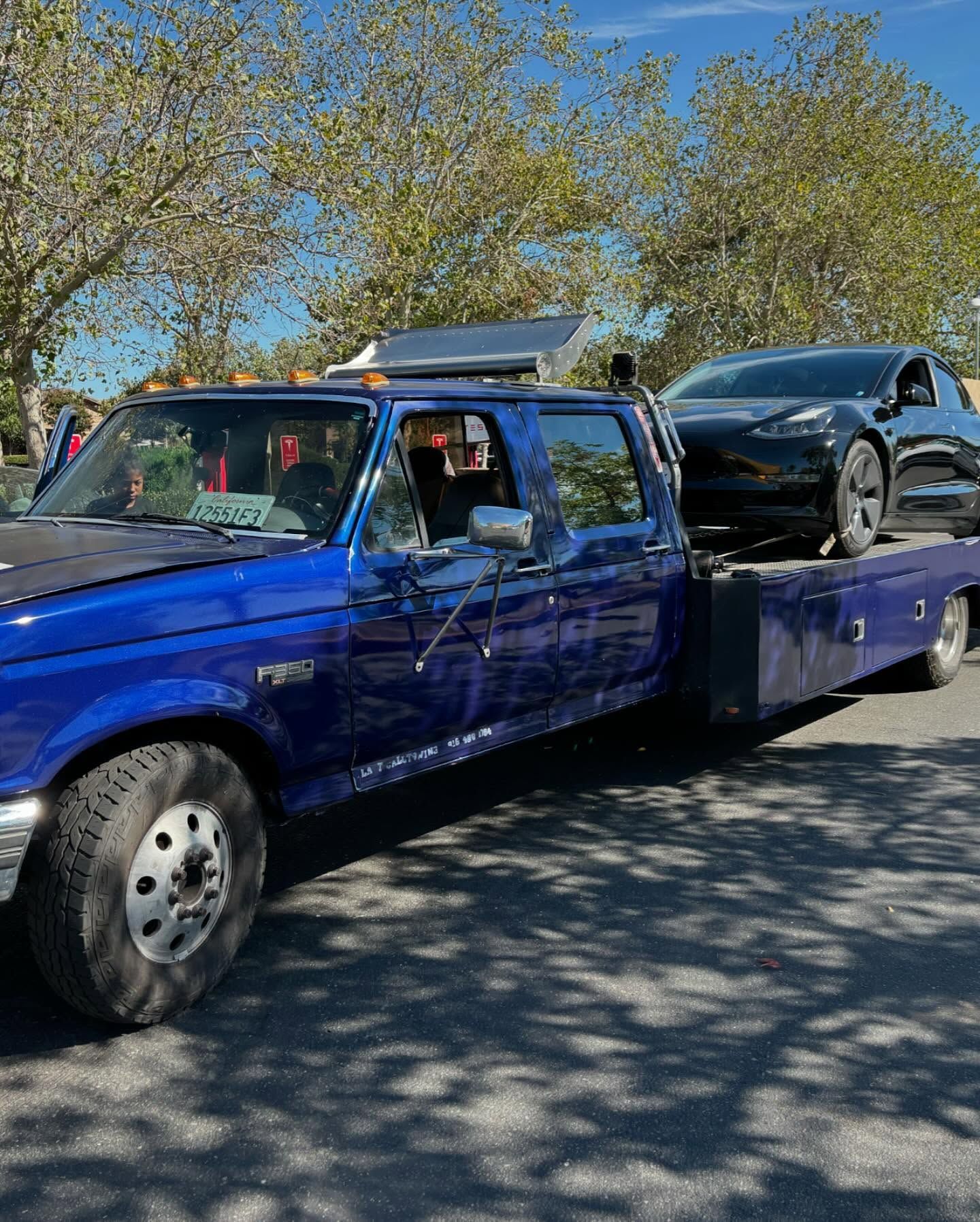 Blue flatbed tow truck carrying a black car on a sunny tree-lined street