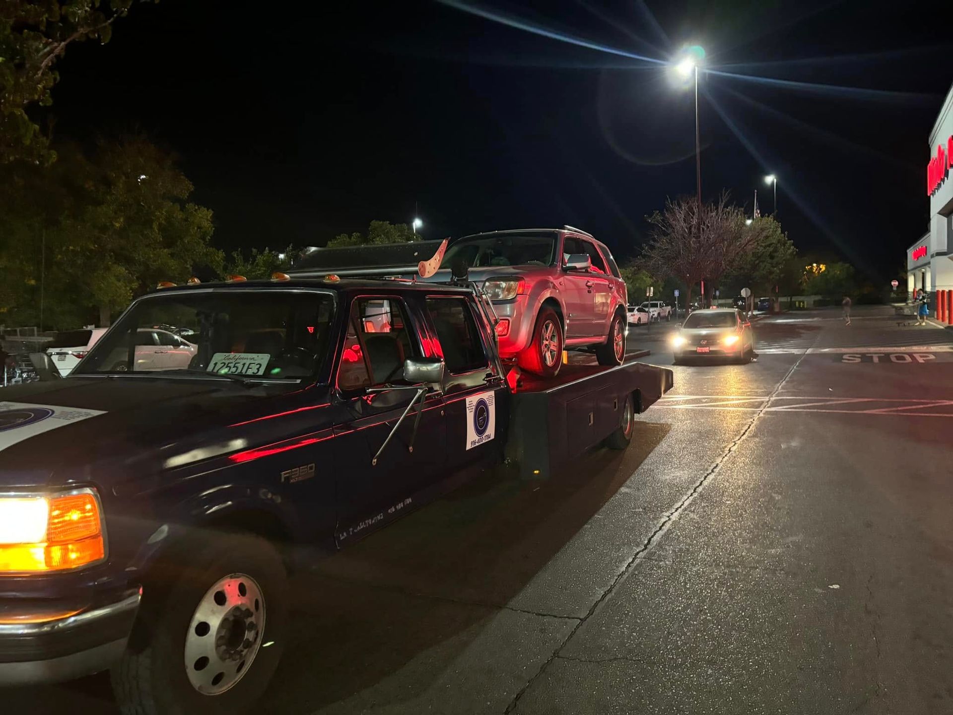 Tow truck carrying a red SUV on a dark street at night, with headlights and store lights visible