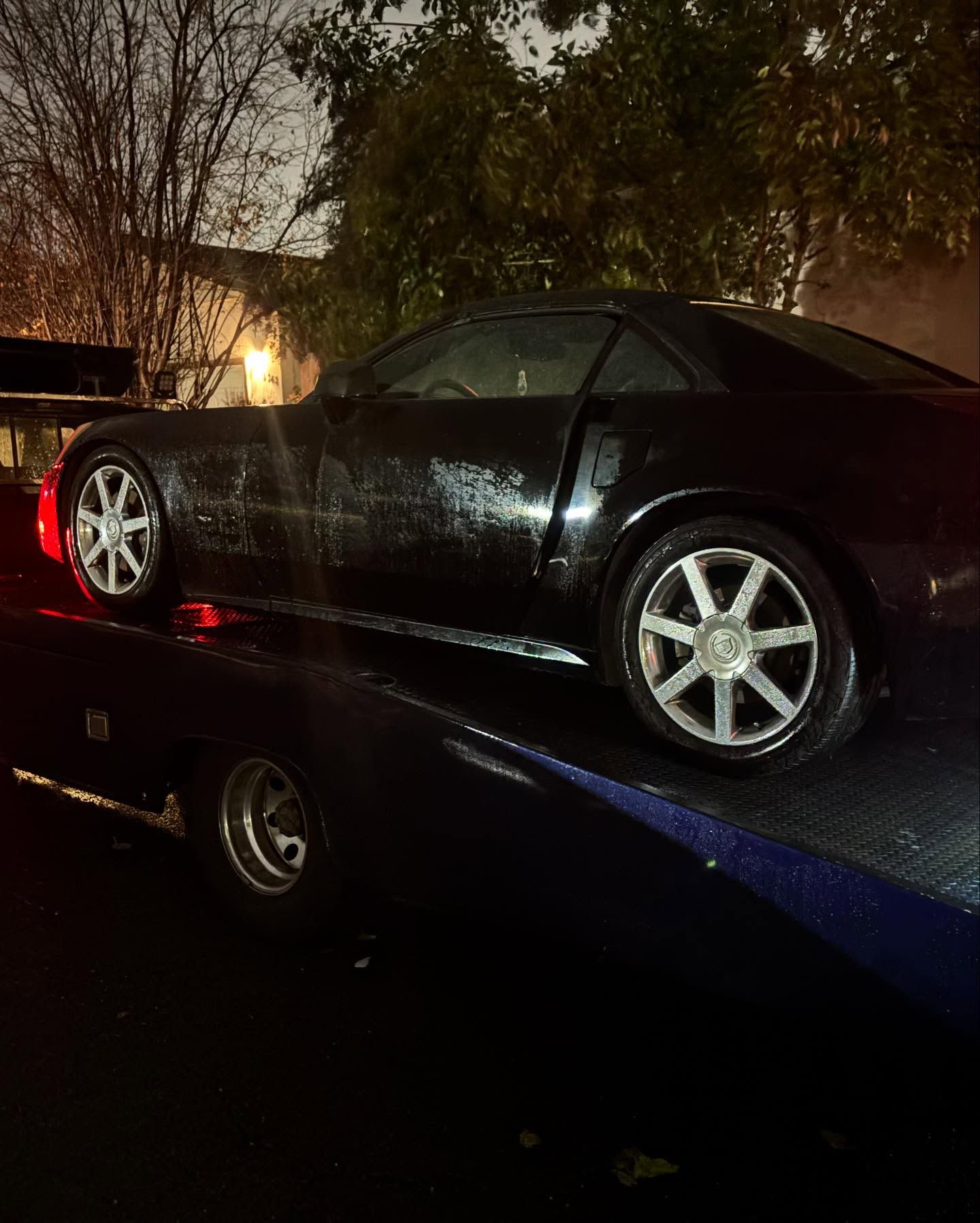 Black sports car on a flatbed tow truck at night, wet from rain, with trees in the background