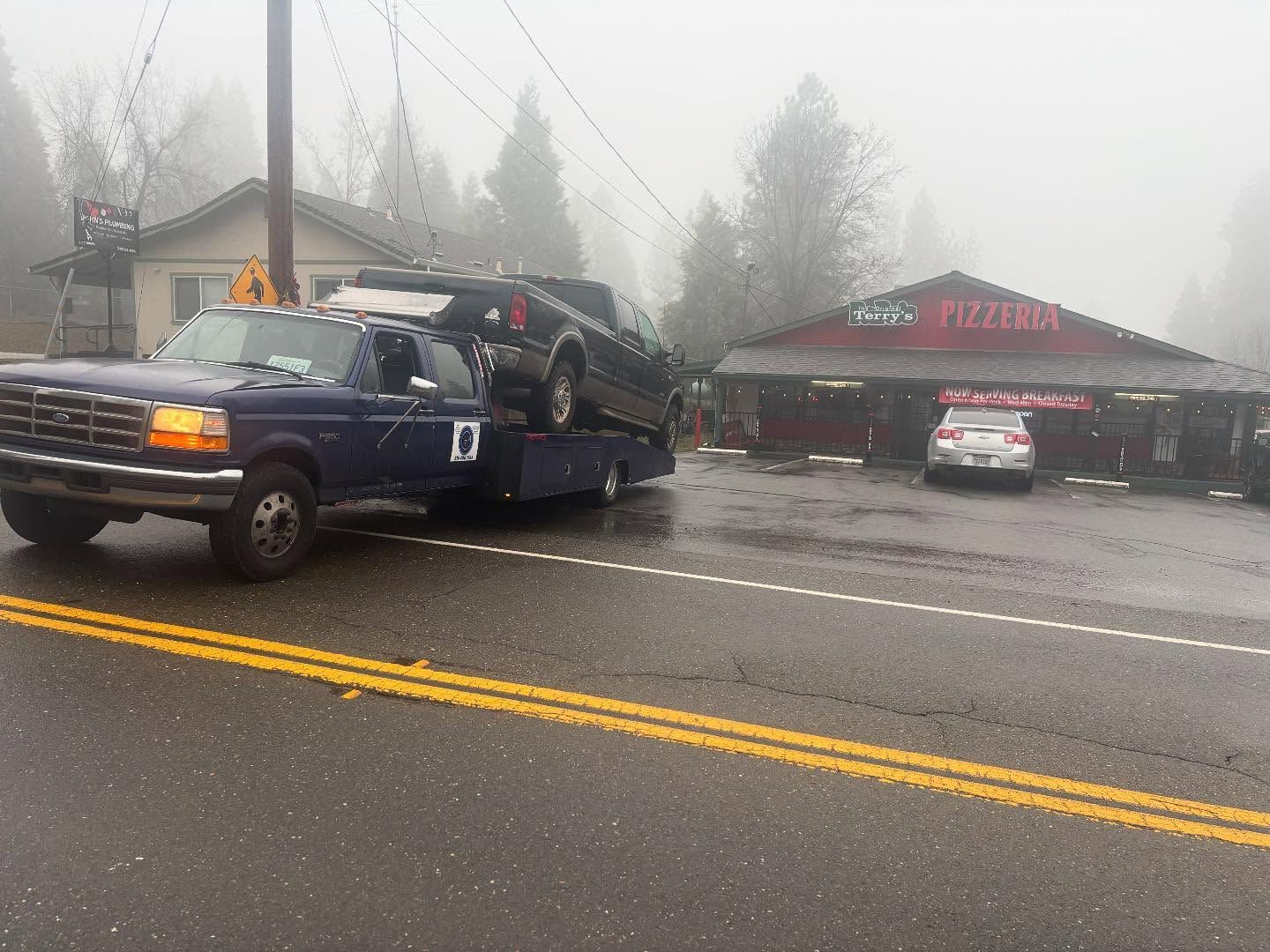Tow truck carrying a dark SUV on a rainy road in front of a building with red awnings