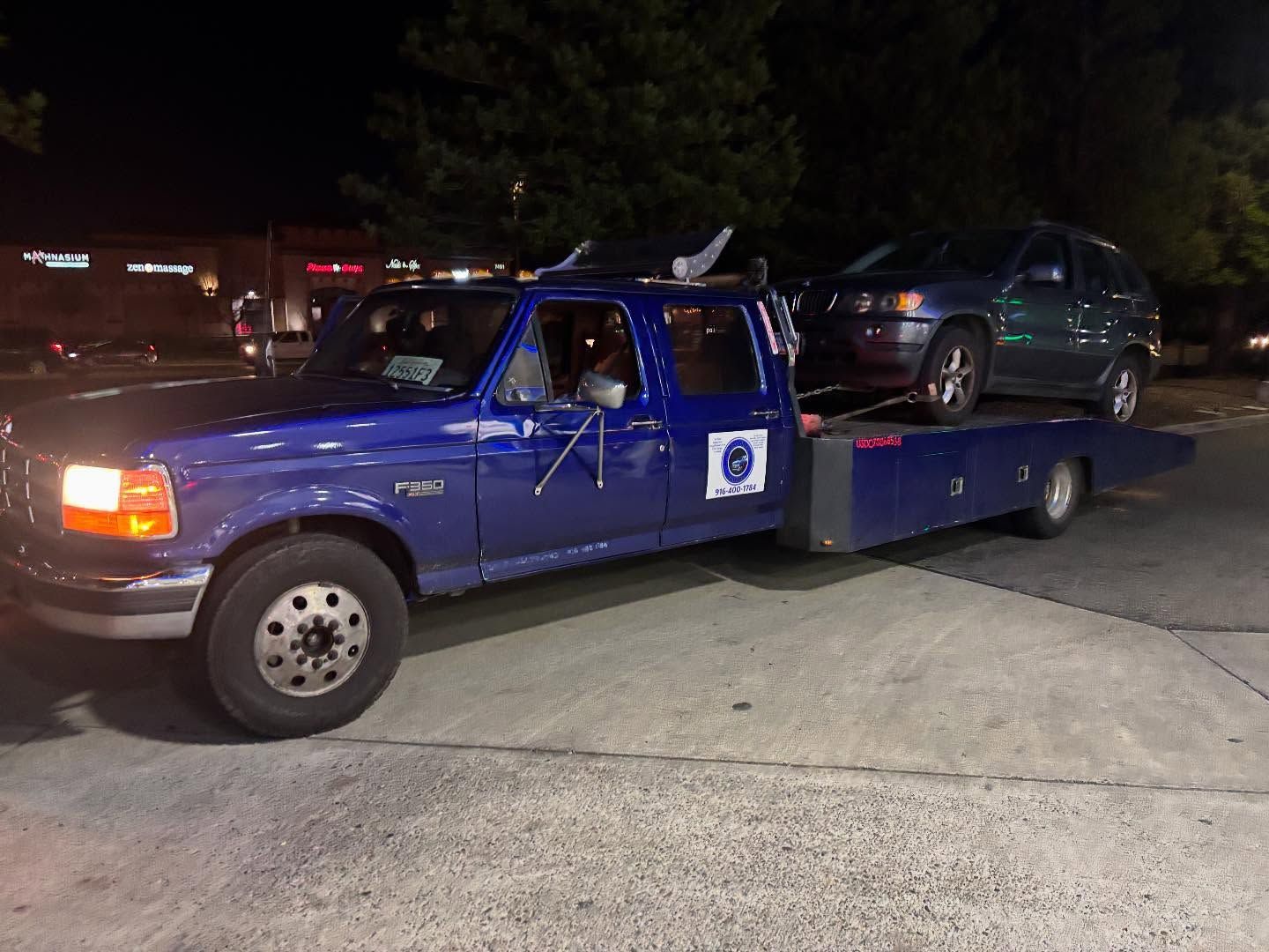 Blue tow truck carrying a dark SUV at night in a parking lot