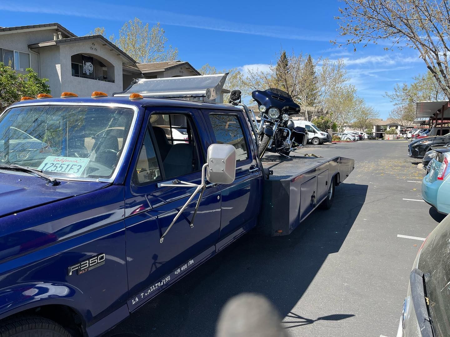 Blue tow truck hauling a motorcycle on a flatbed along a sunny residential street