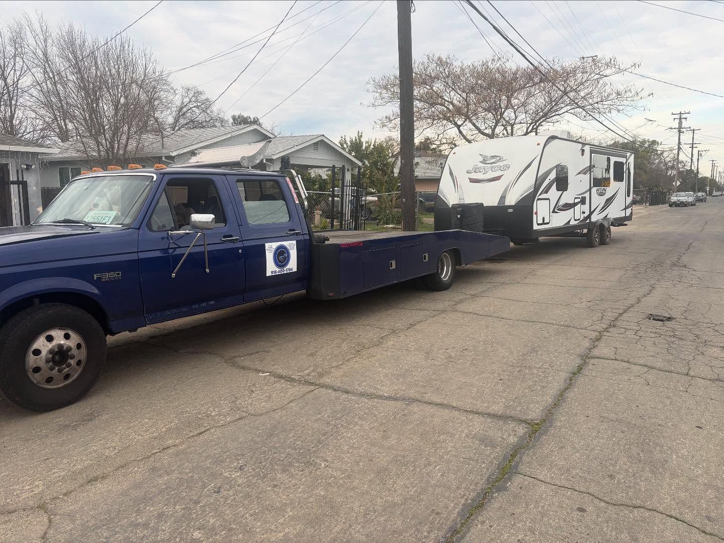 Blue tow truck hauling a white RV on a residential street under overcast skies