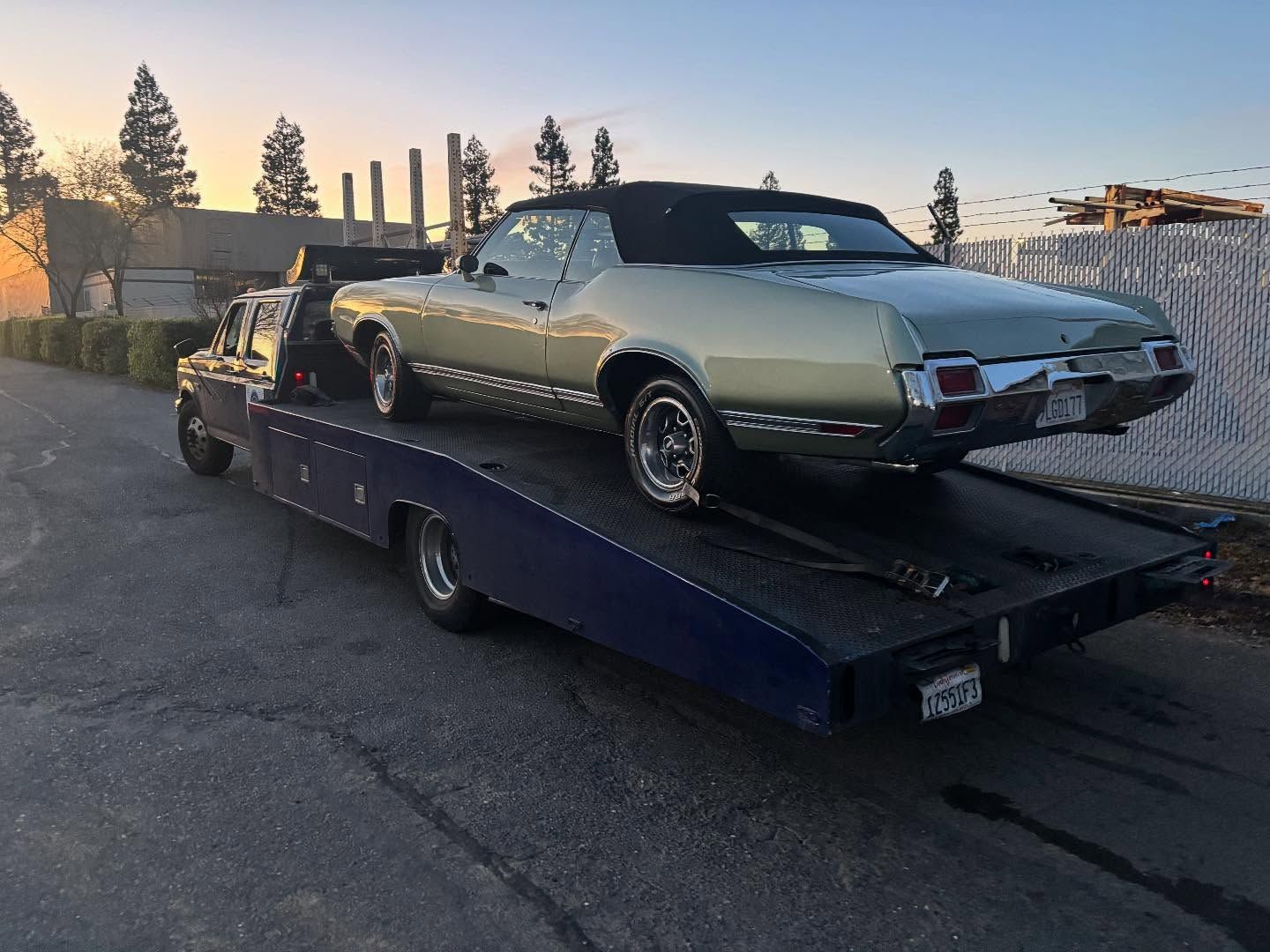 Classic convertible on a flatbed tow truck at sunset, parked in a lot.