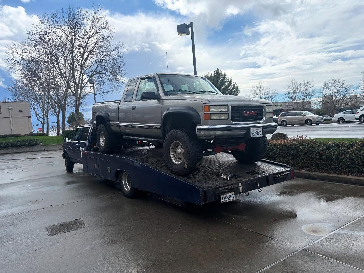 Silver pickup truck on a flatbed tow truck in a wet parking lot under a cloudy sky