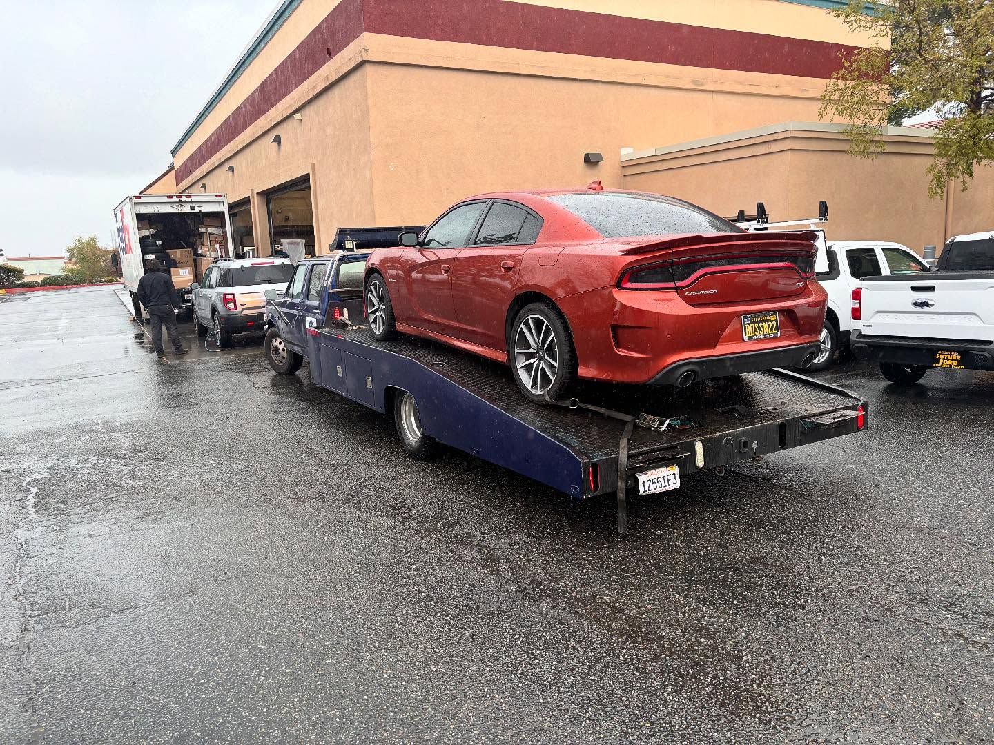 Red sports car on a flatbed tow truck in a wet parking lot outside a tan building.
