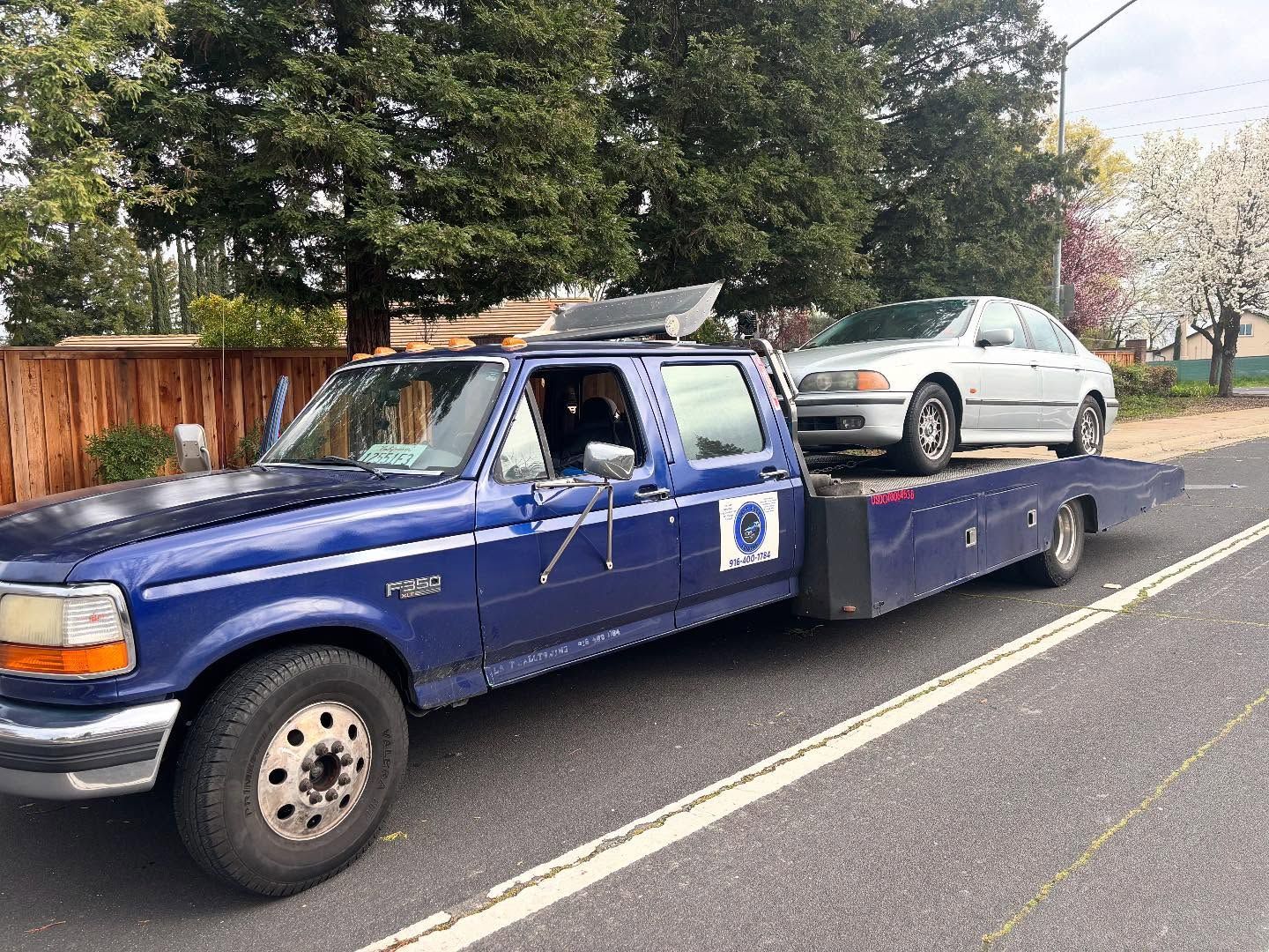 Blue tow truck hauling a silver car on a roadside lane, with trees and a wooden fence in the background