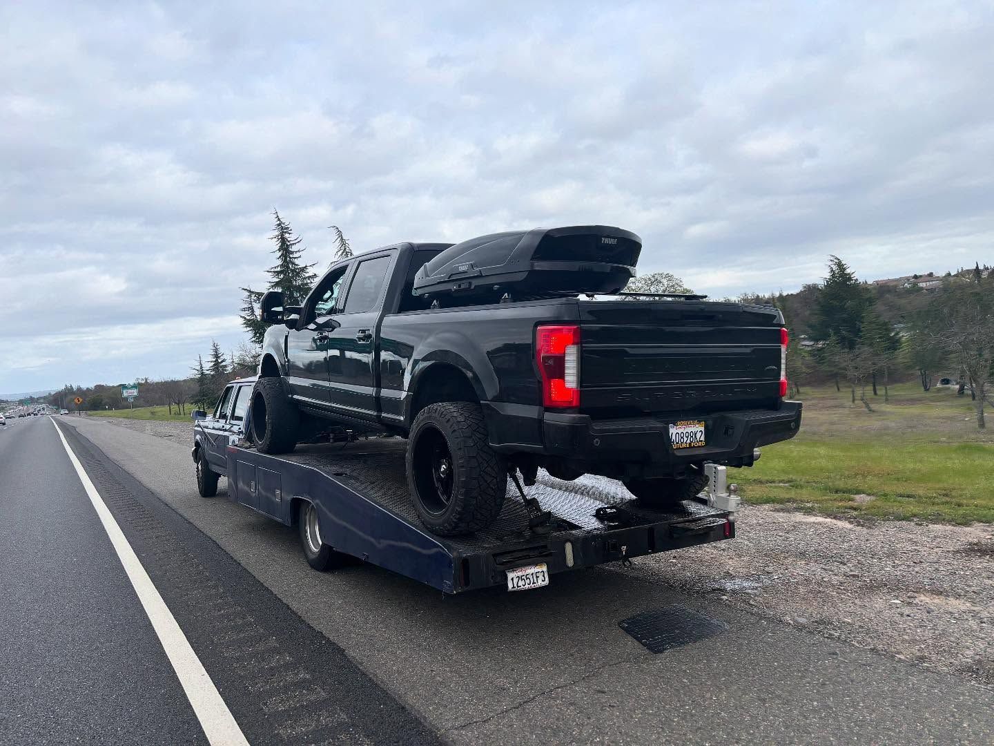 Black pickup truck loaded on a blue flatbed trailer on a roadside under cloudy skies.