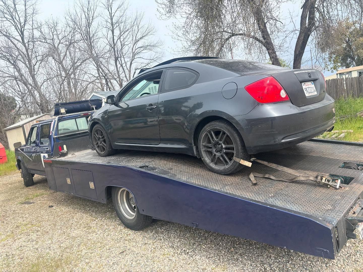 Dark gray coupe strapped on a flatbed tow truck in a wooded outdoor area
