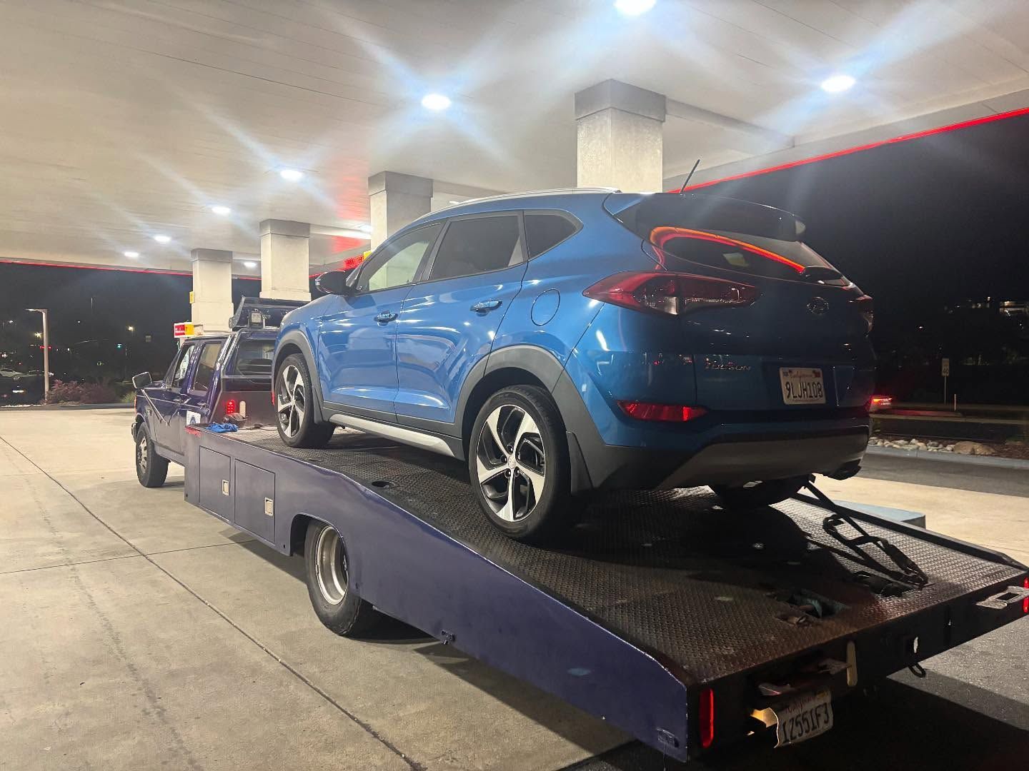 Blue SUV on a car hauler trailer at a gas station at night, with another vehicle in front.