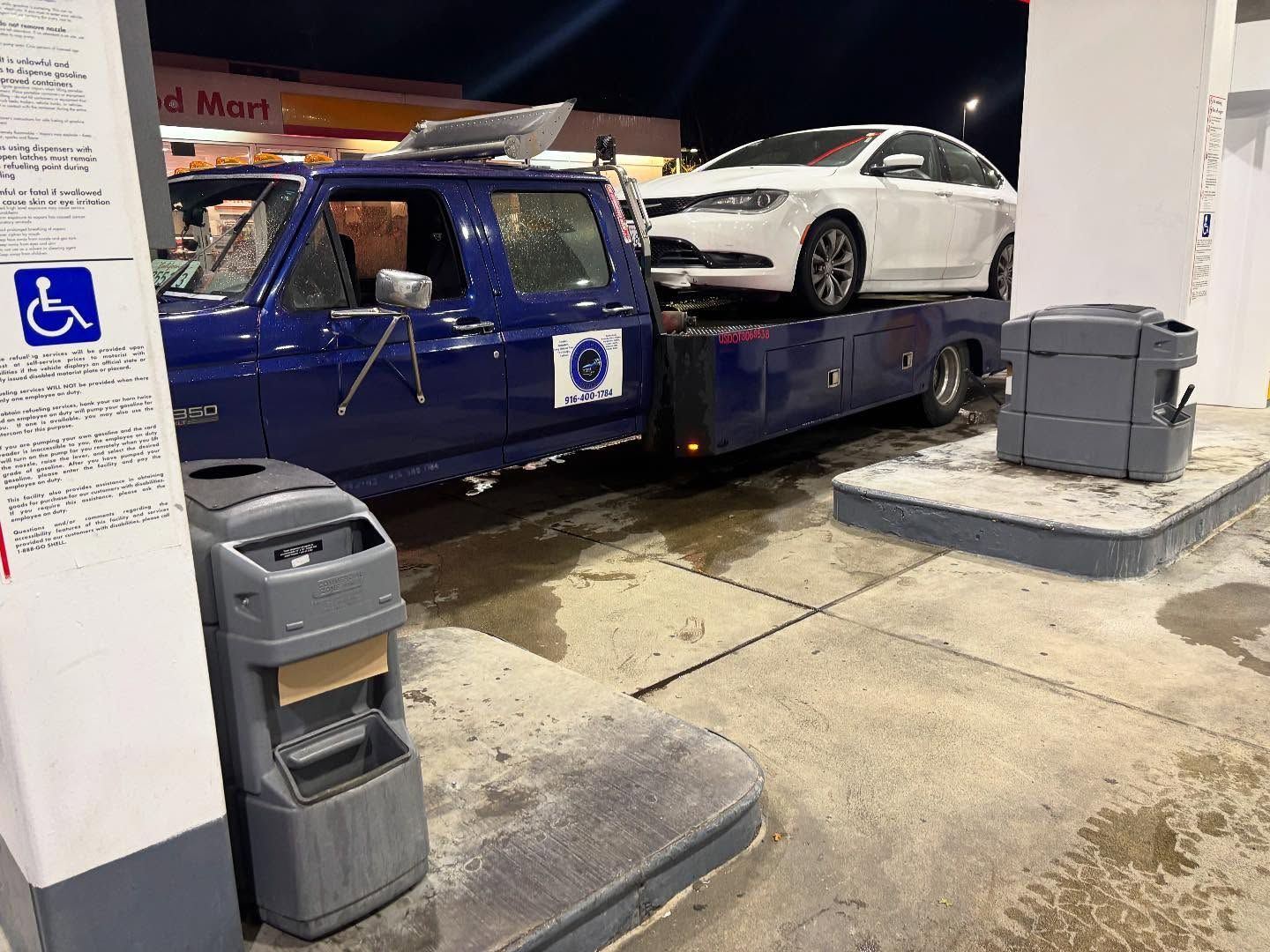 Blue tow truck loading a white sedan at a gas station at night