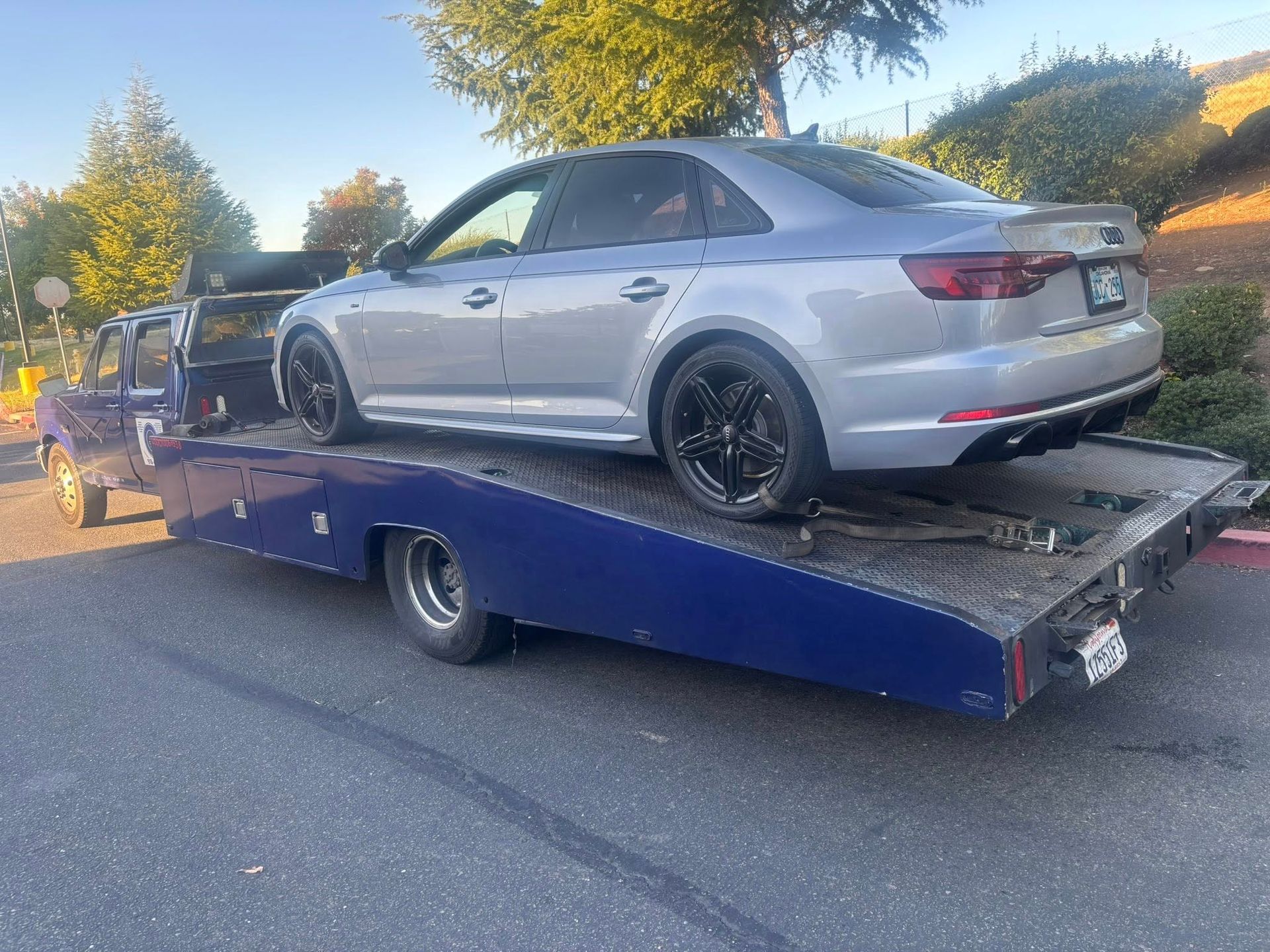 Silver sedan on a blue tow trailer on a road, with trees and daylight in the background