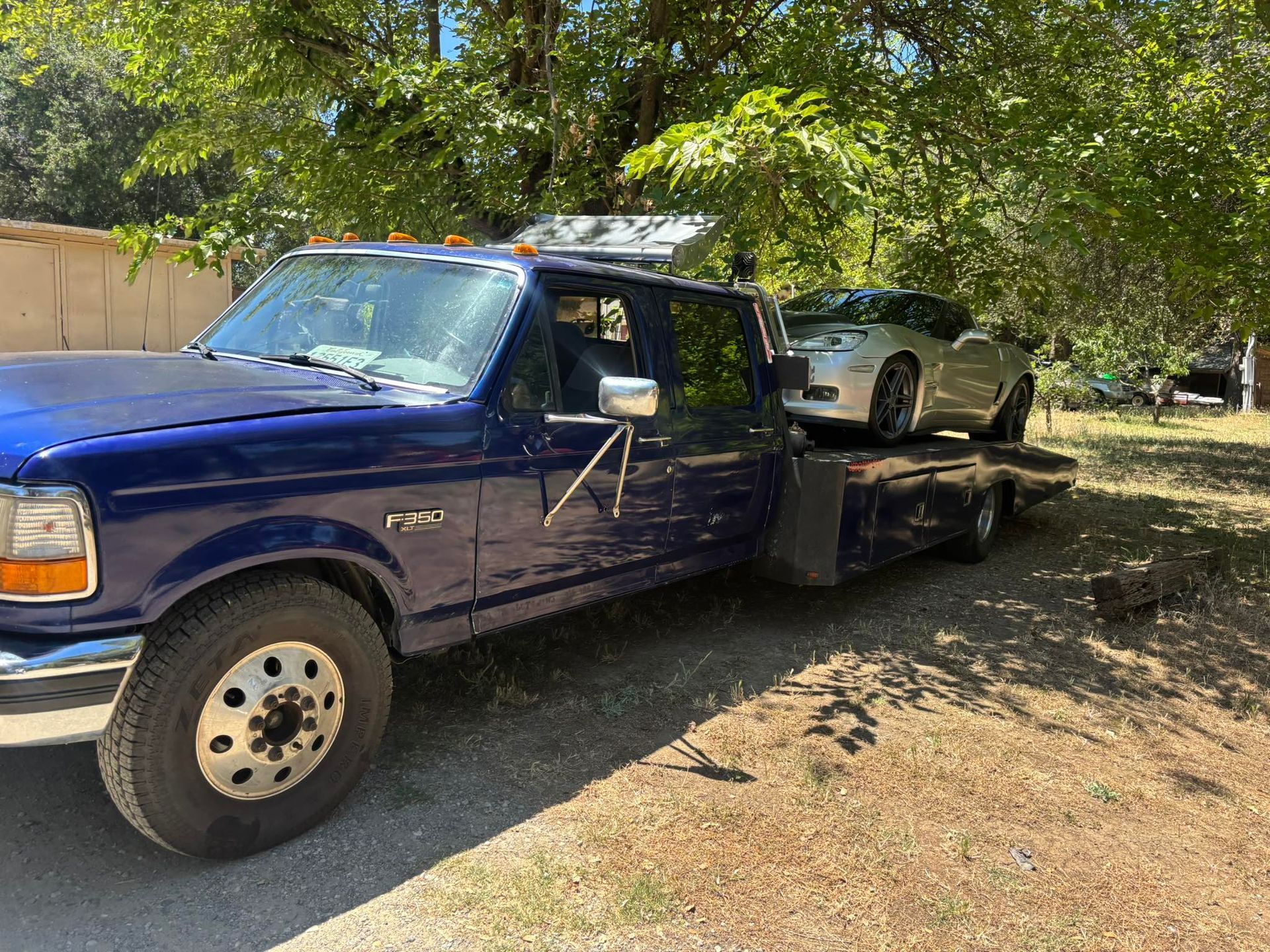 Blue tow truck carrying a silver car on a flatbed in a shaded yard