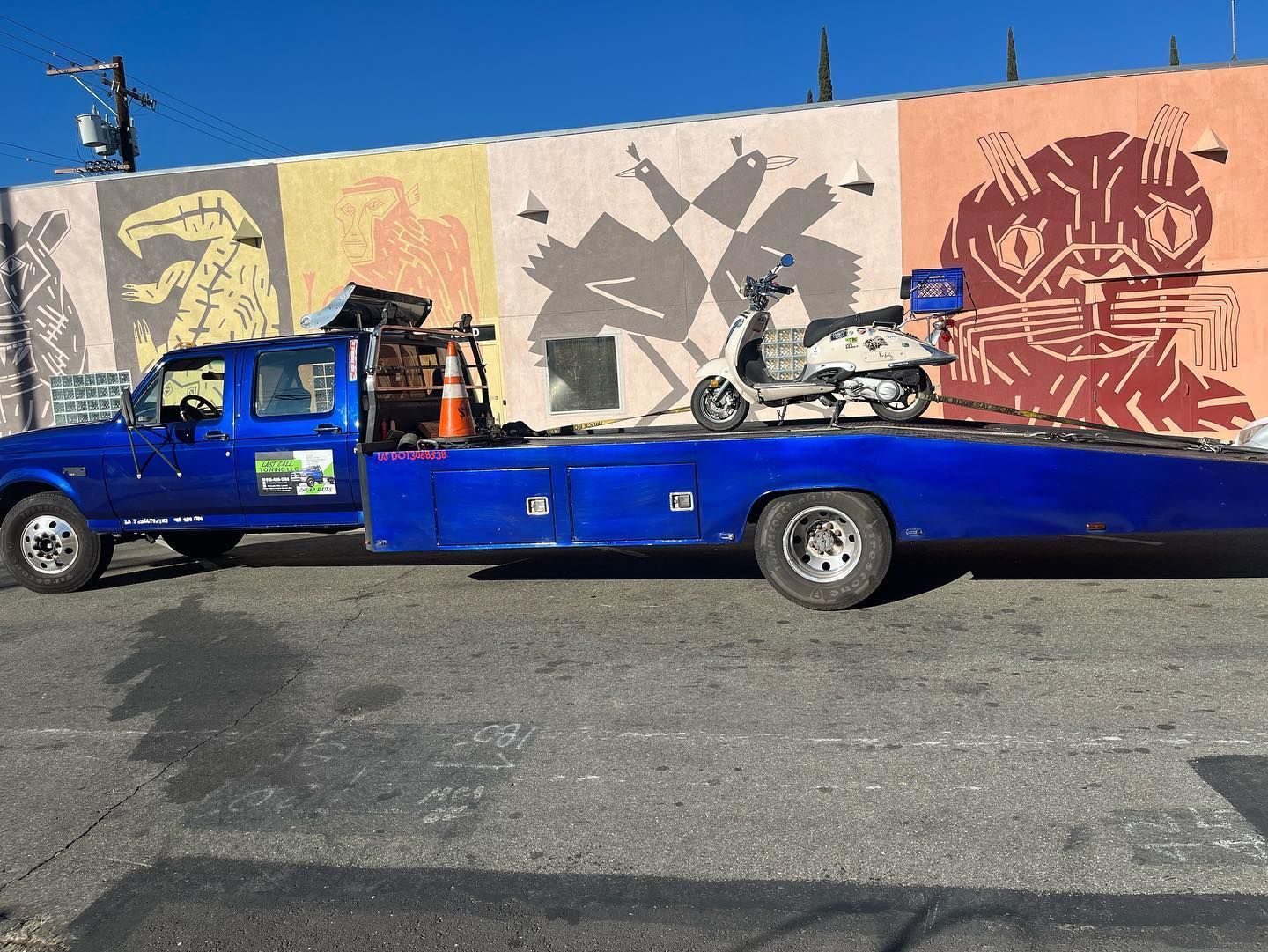 Blue flatbed truck parked by a mural-covered wall with a motorcycle loaded on the bed