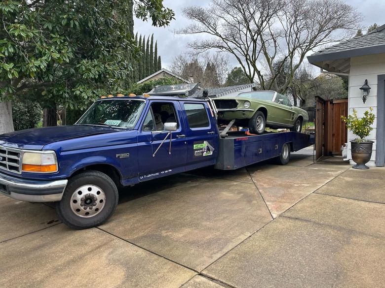 Blue tow truck carrying a green car in a driveway beside a house