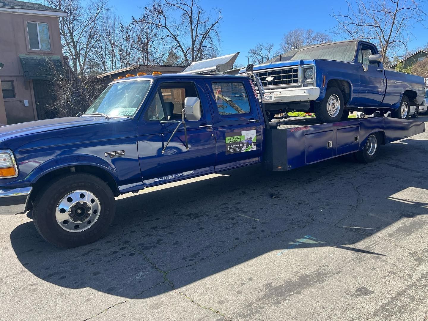 Blue flatbed tow truck carrying a blue pickup on a sunny residential street.