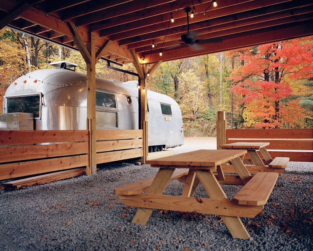 An airstream trailer is parked under a covered area with picnic tables