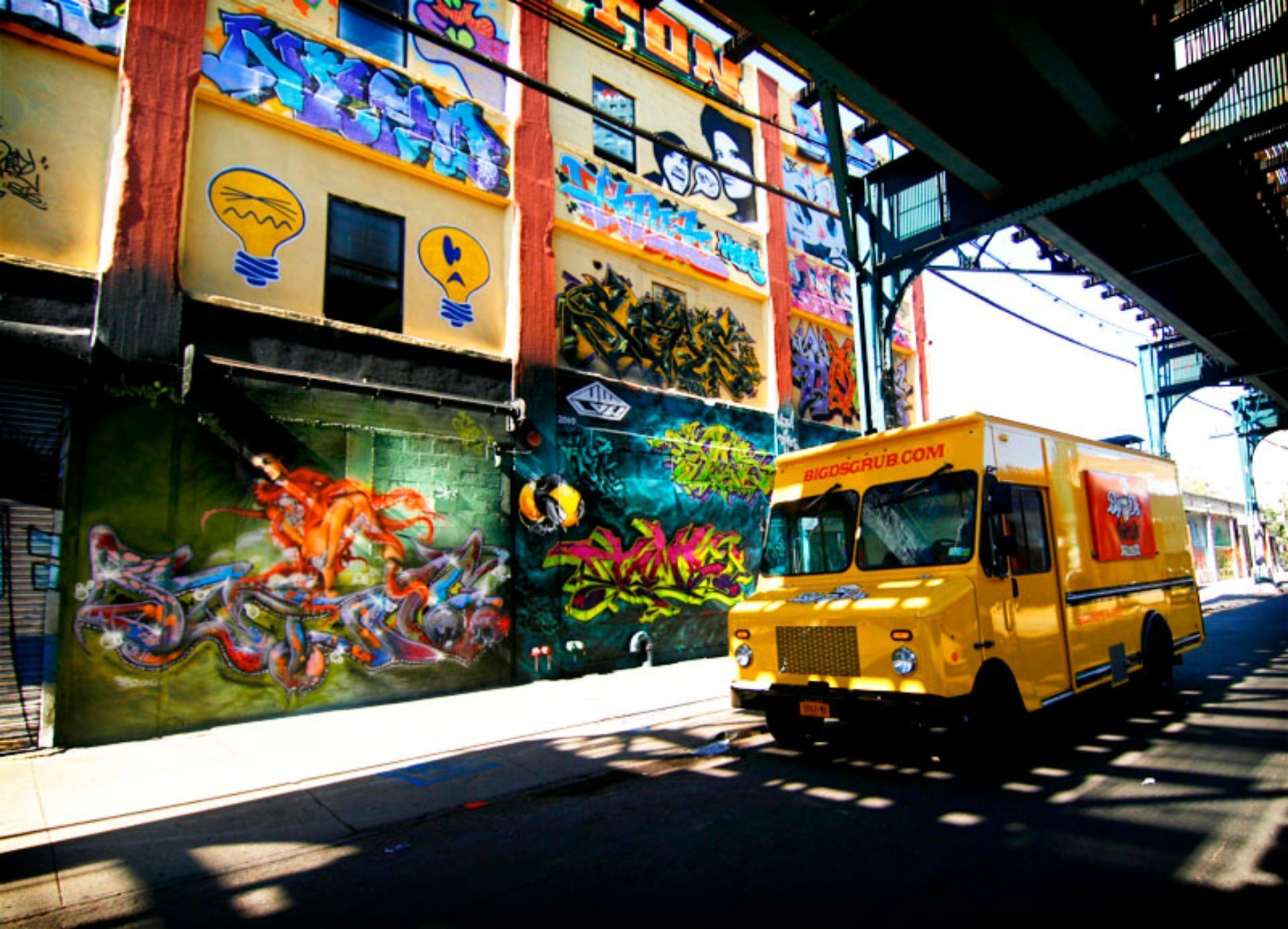 A yellow food truck is driving under a bridge in front of a building with graffiti on it