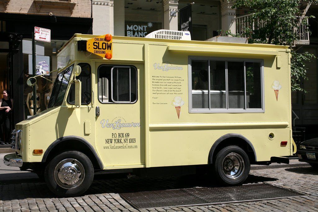 A yellow ice cream truck is parked on a cobblestone street