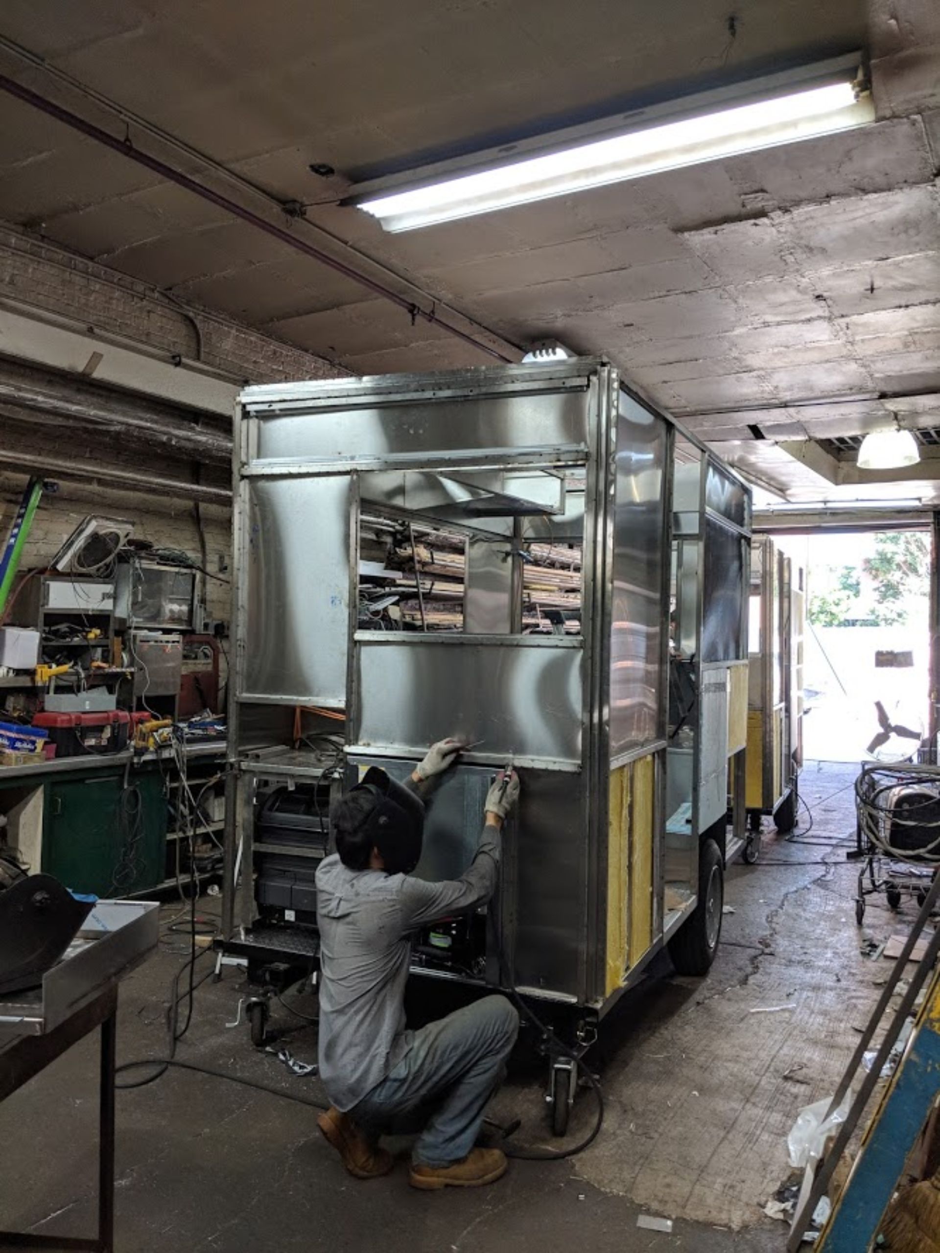 A man is working on a stainless steel food truck in a garage.