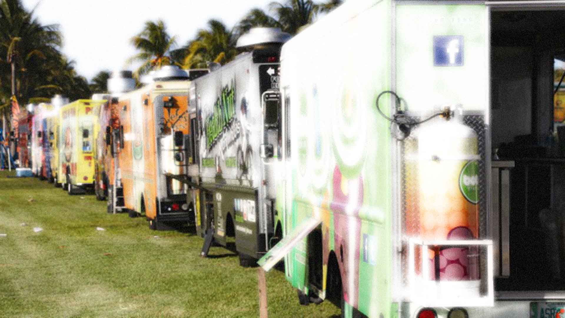 A row of food trucks are parked in a grassy field.