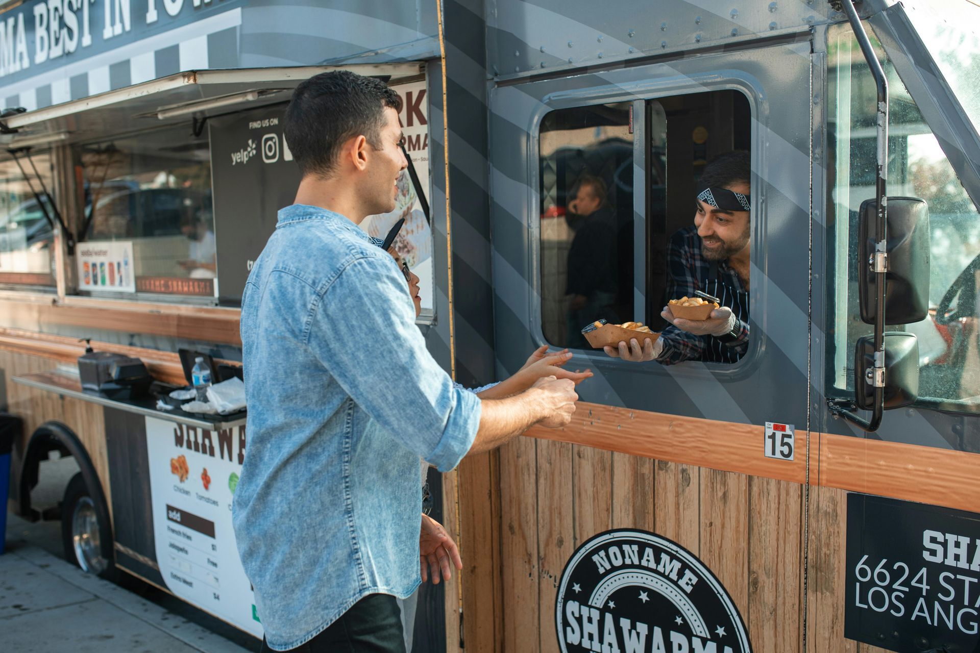 Man receiving food from a food truck window. Two men are visible. The truck has a wooden front, and there is a sunny exterior.