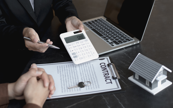 Hands reviewing a contract with calculator, laptop, and house model on a desk