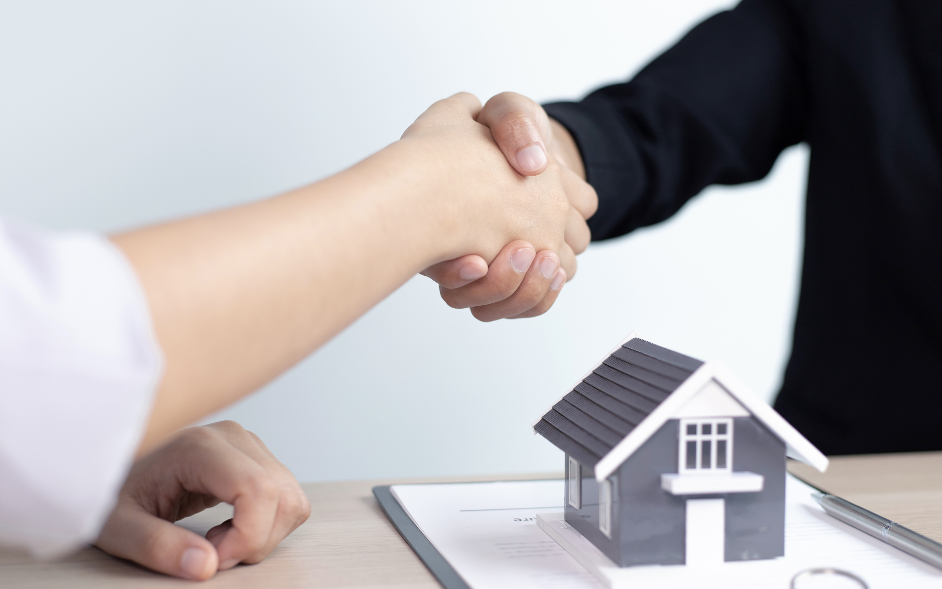 Handshake over a desk beside a small house model and paperwork, suggesting a real estate deal