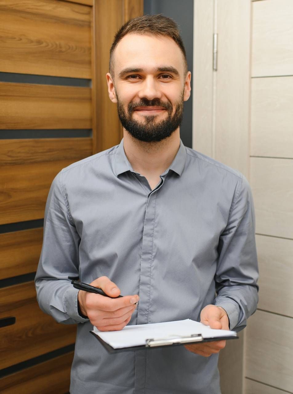 Man standing indoors with a clipboard and pen, wearing a gray shirt and smiling near a wooden door.