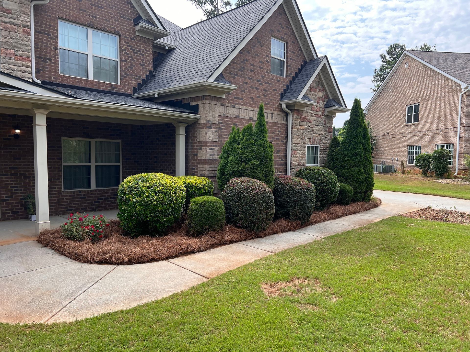 Brick house front yard with trimmed shrubs, driveway, and covered porch.