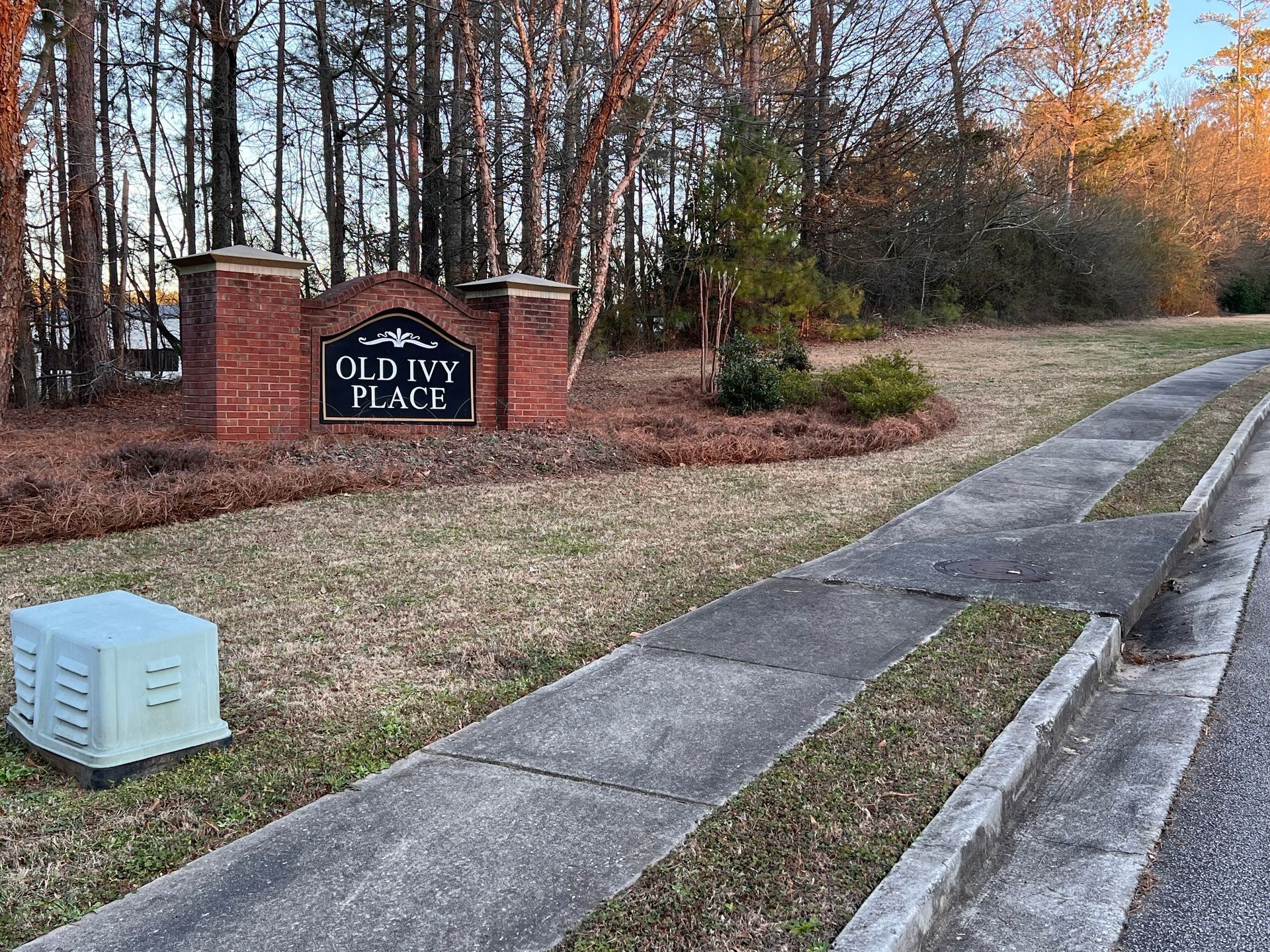 Brick neighborhood entrance sign by a sidewalk and road, with trees and a utility box nearby.