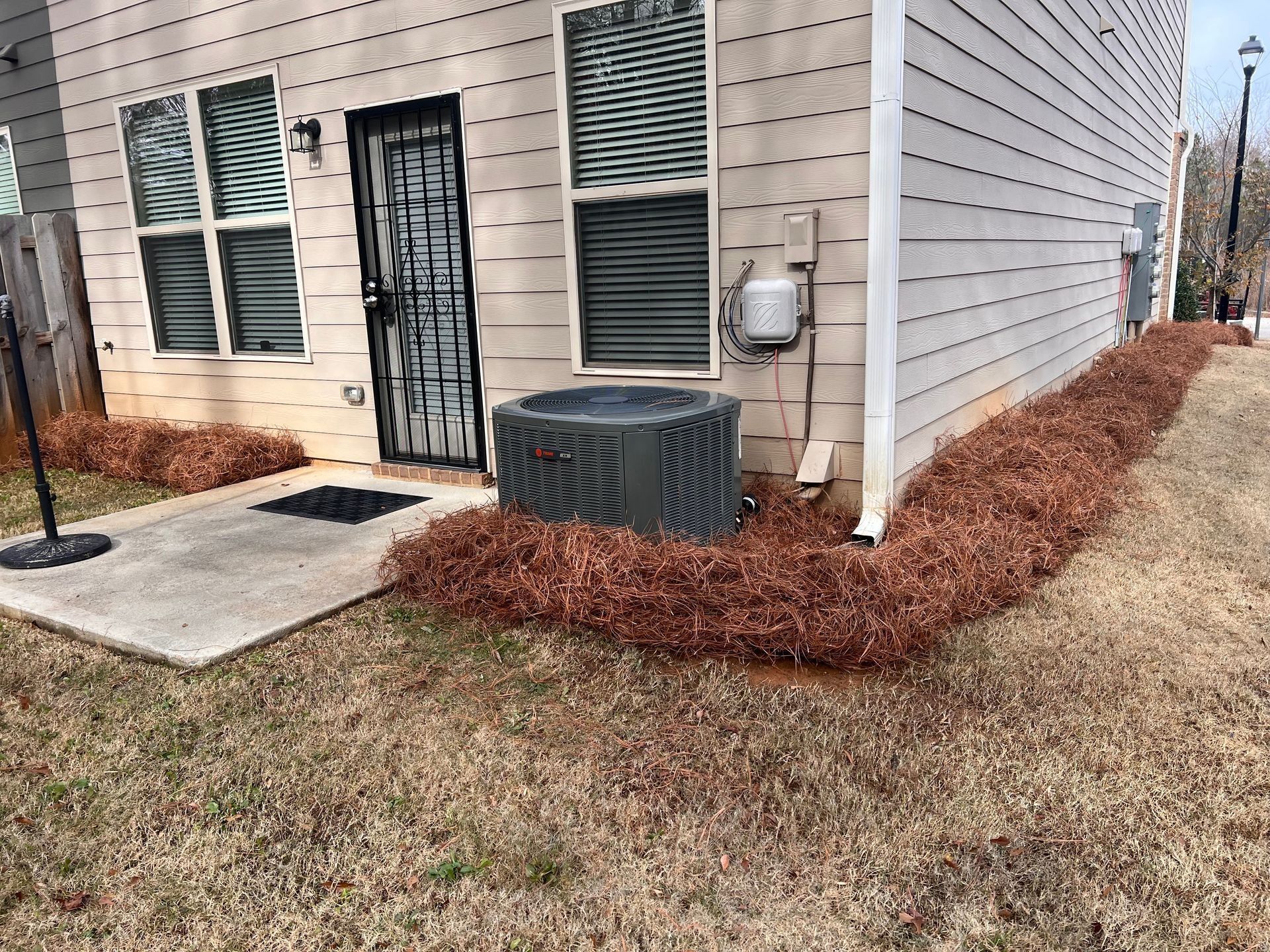 Front entrance of a house with windows, a black door, HVAC unit, and mulch beds along the siding.