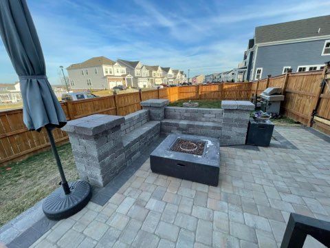 Backyard patio with stone fire pit, seating, and wooden fence under a blue sky
