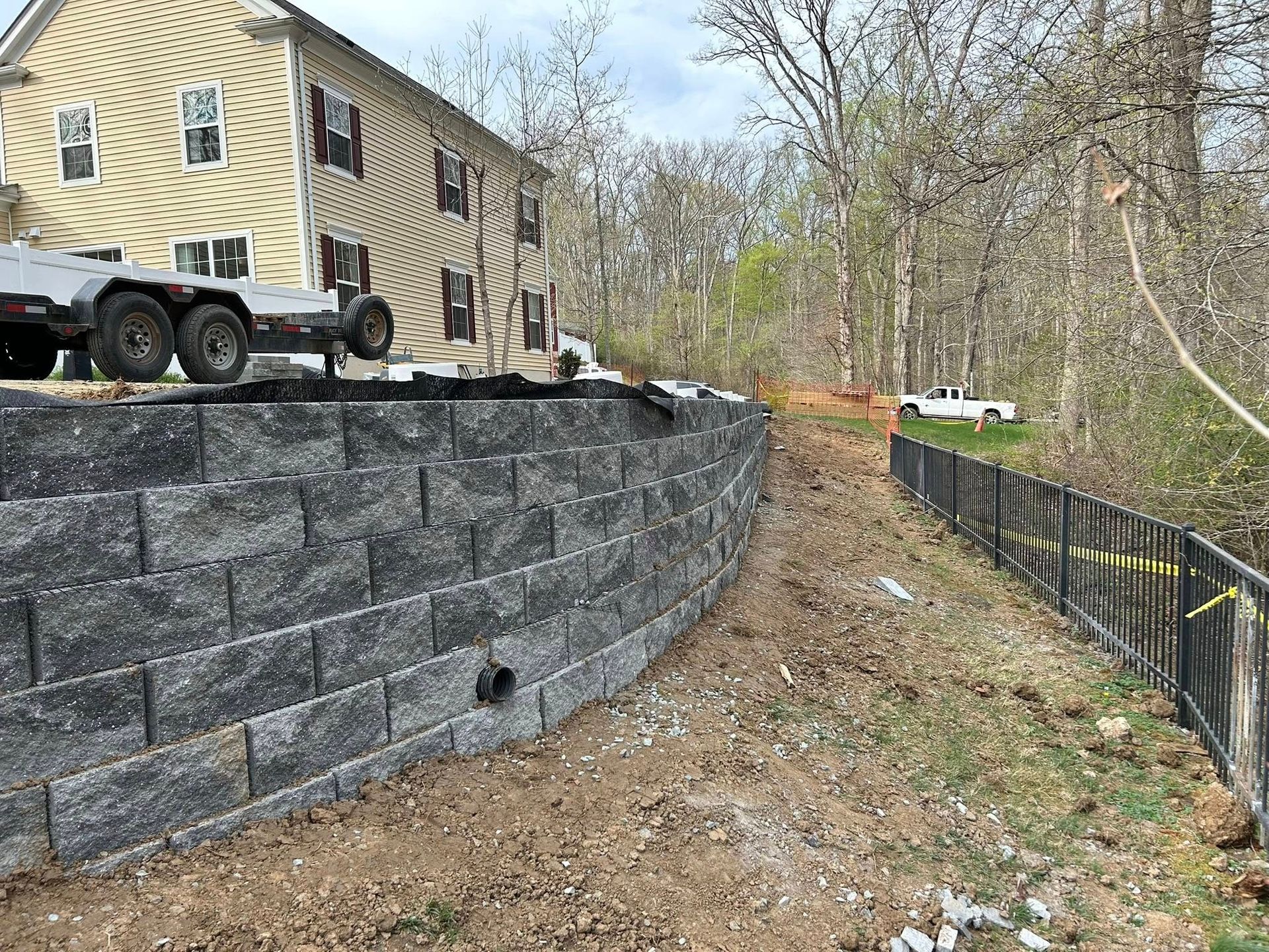 Stone retaining wall beside a tan building on a sloped construction site with an excavator and erosion fencing