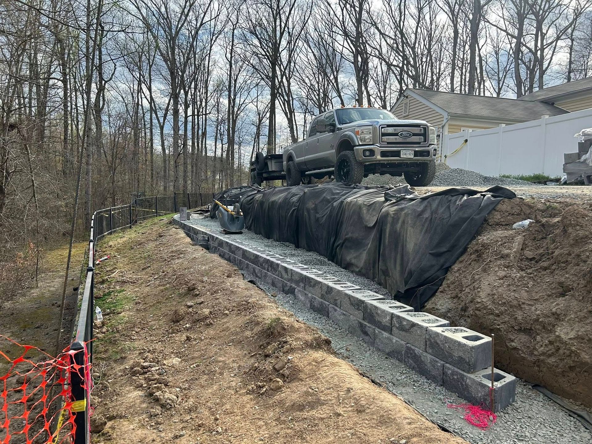 Construction trucks on a sloped dirt embankment beside a black retaining wall and wooded area.