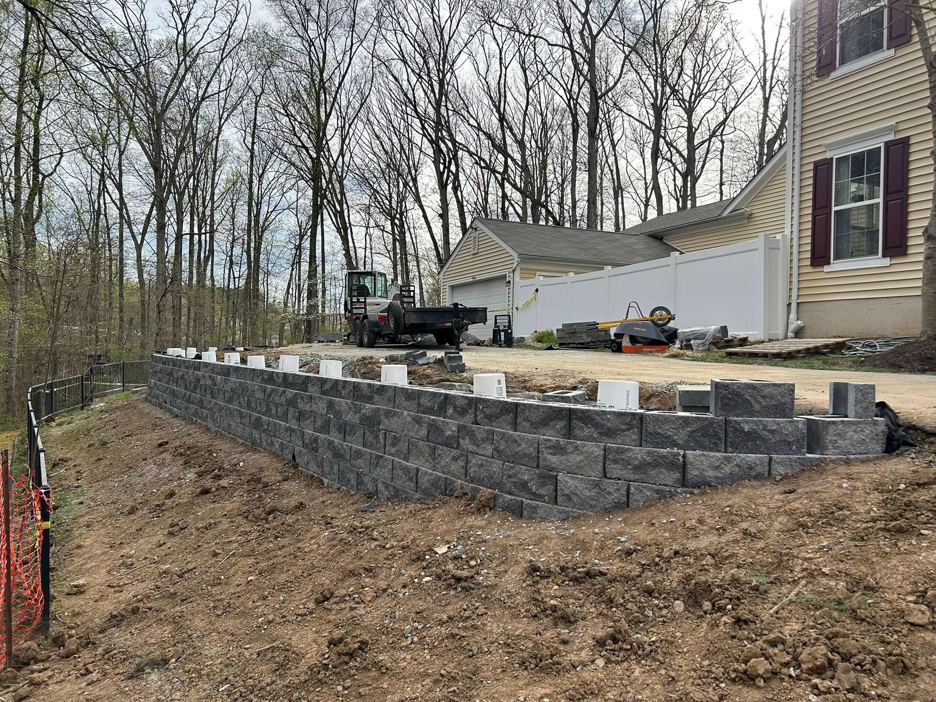 Stone retaining wall terraced beside a house on a sloped, wooded yard under construction