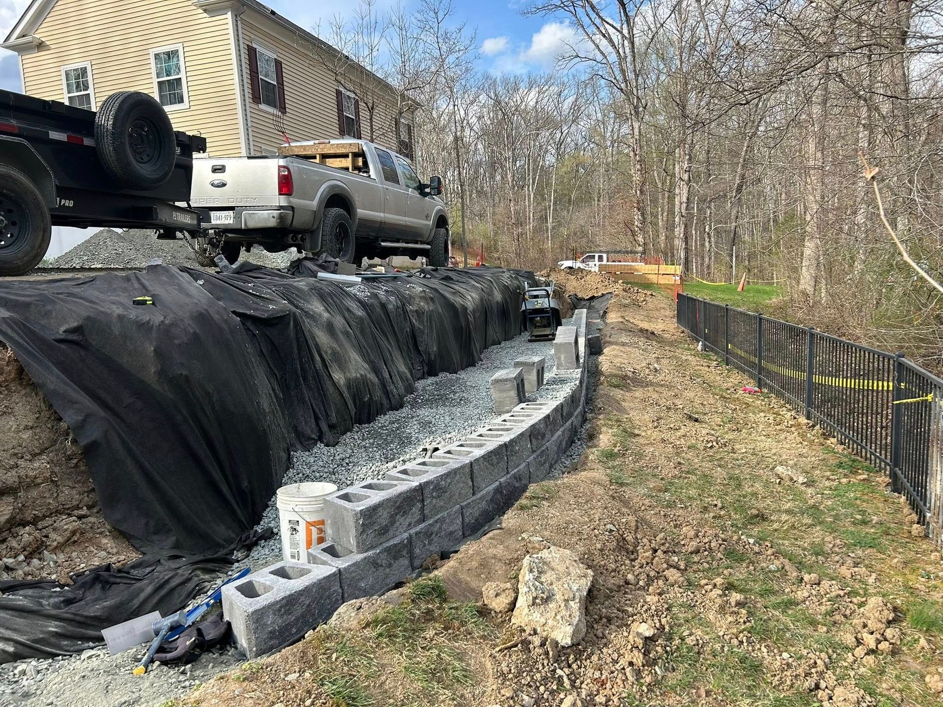 Curved stone retaining wall under construction beside a house, with a pickup truck and black landscaping fabric nearby