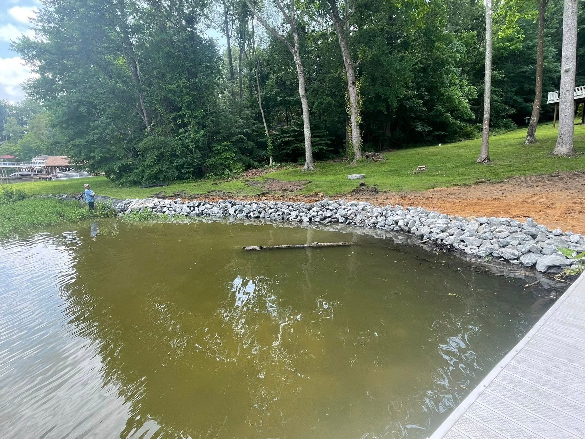 Small pond with muddy water, rocky shoreline, and trees along a grassy bank.