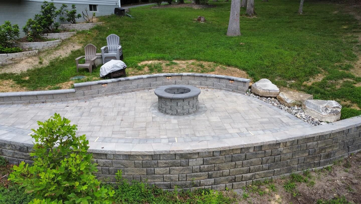 Stone patio fire pit in a grassy backyard with a curved retaining wall and surrounding rocks
