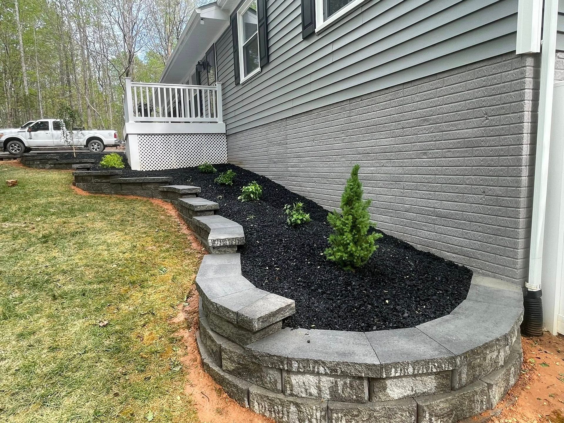Curved stone flower bed with black mulch and small shrubs beside a gray house