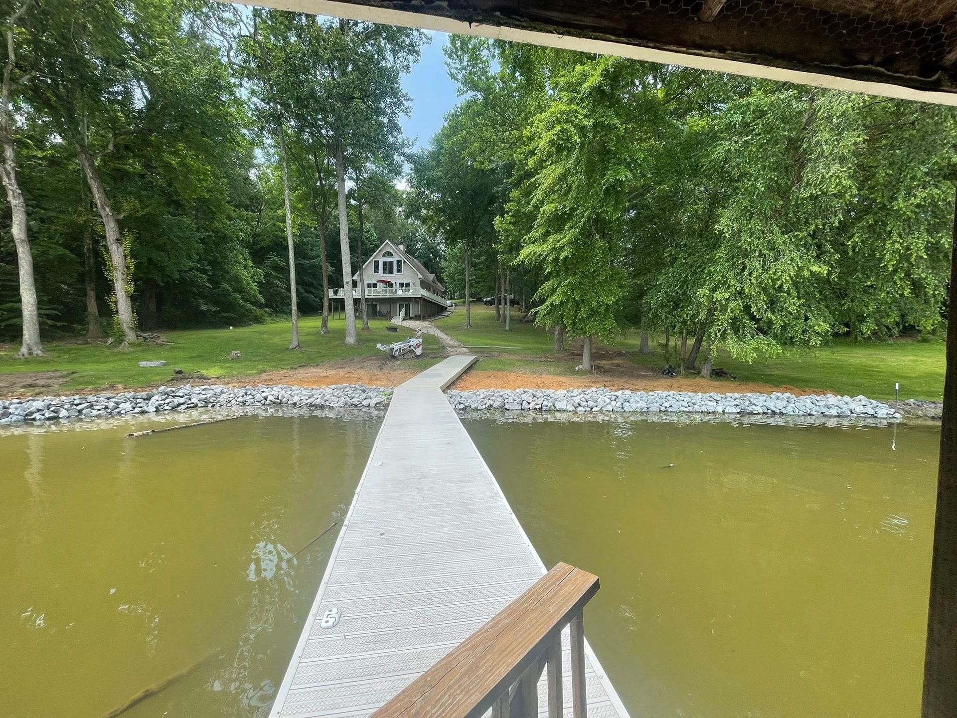Wooden dock over green water leading to a small lakeside cabin surrounded by trees