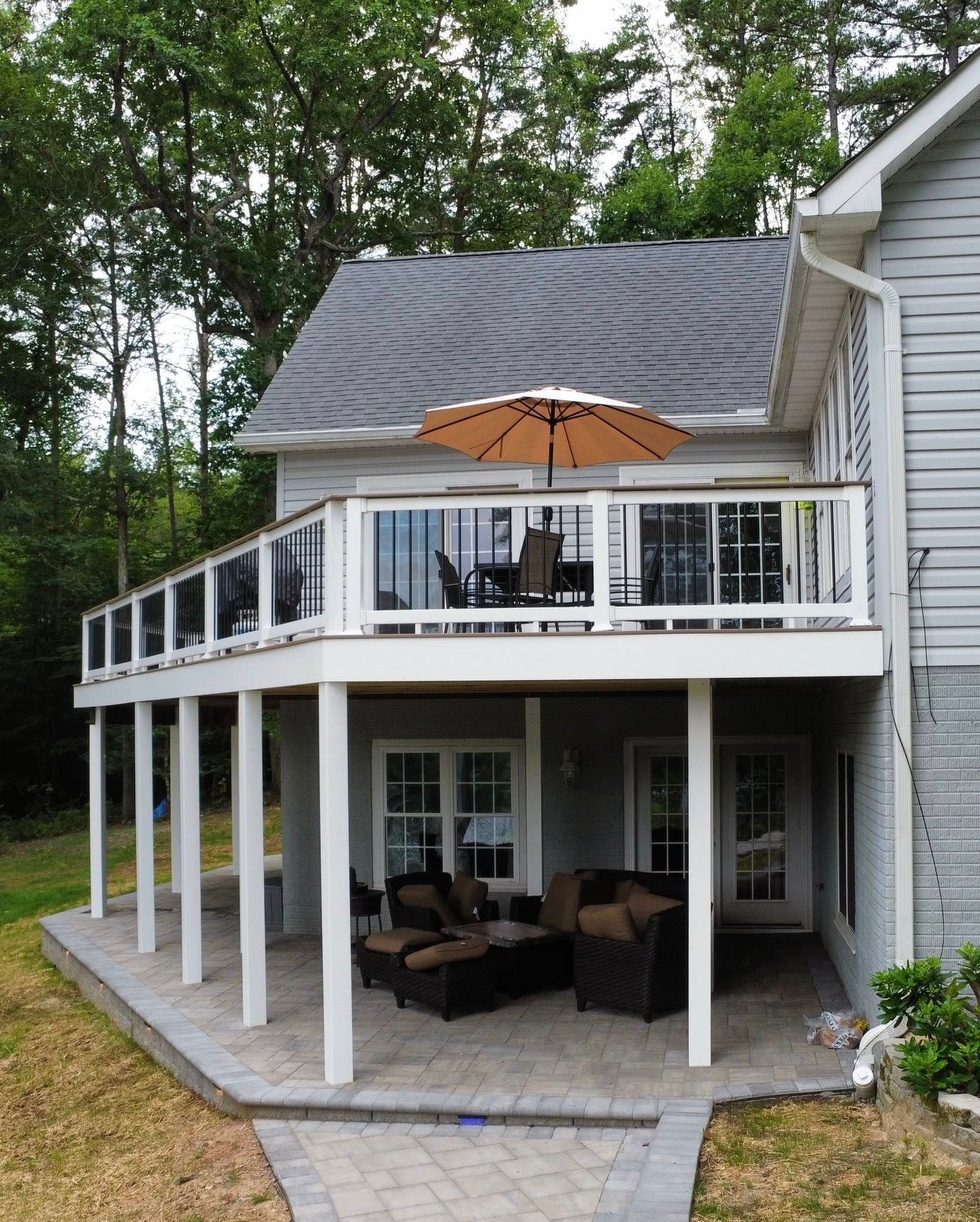 Two-story house with a white-railed deck, patio seating below, and a large umbrella on the upper level.