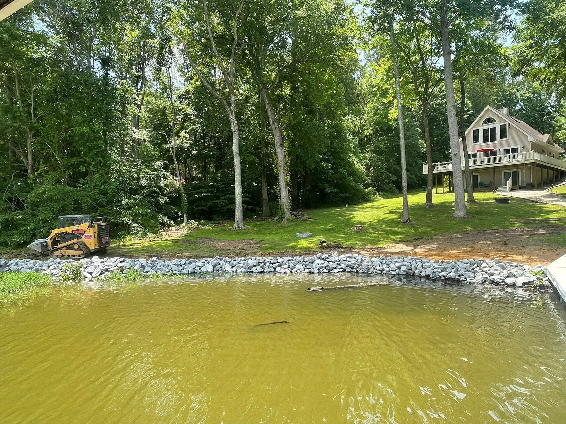 Shoreline with green water, rock embankment, wooded trees, and a house by the lake