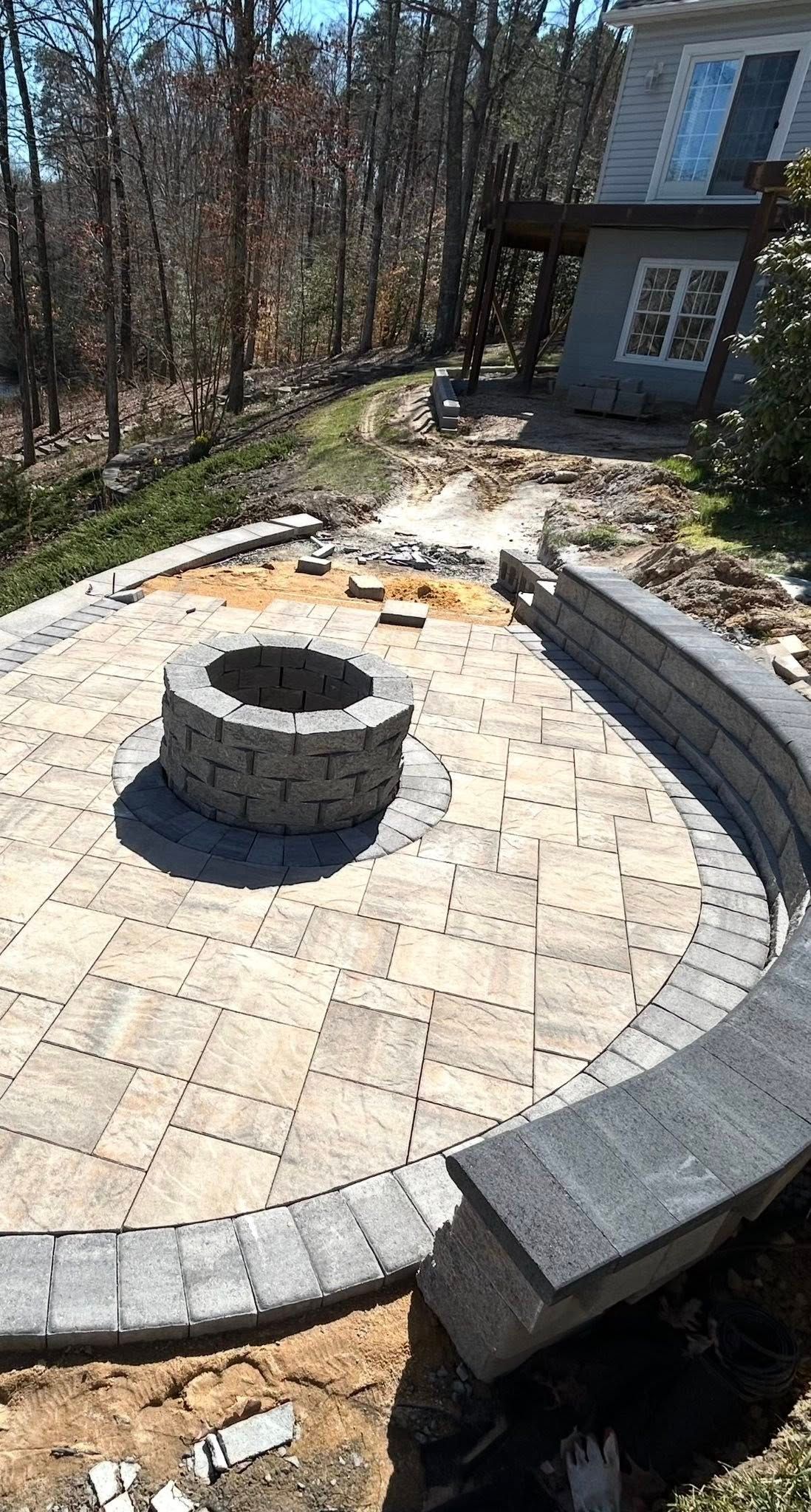 Stone patio with a circular fire pit and curved bench beside a house, surrounded by trees.