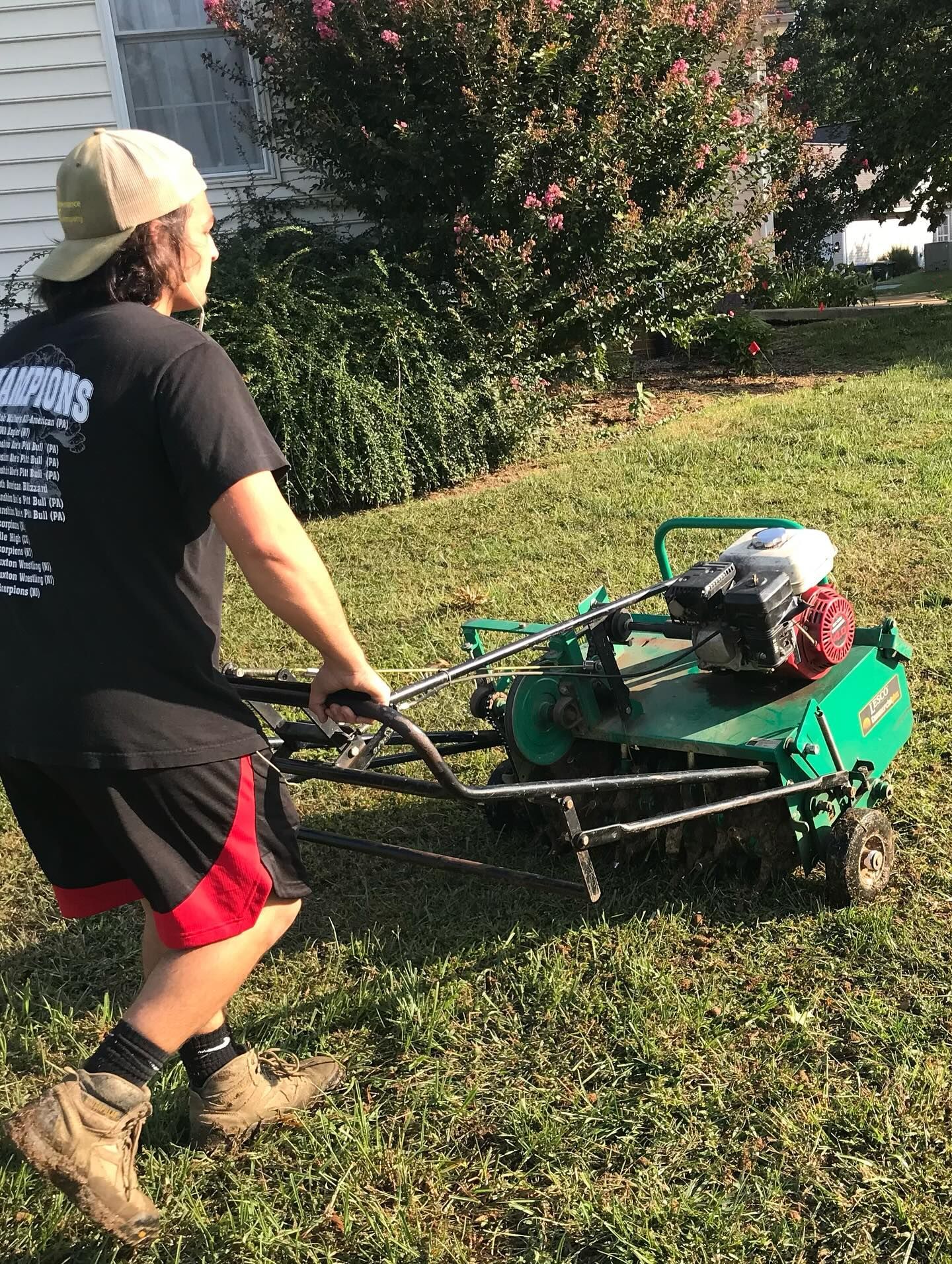 Person operating a green lawn mower on a grassy lawn near shrubs and a house