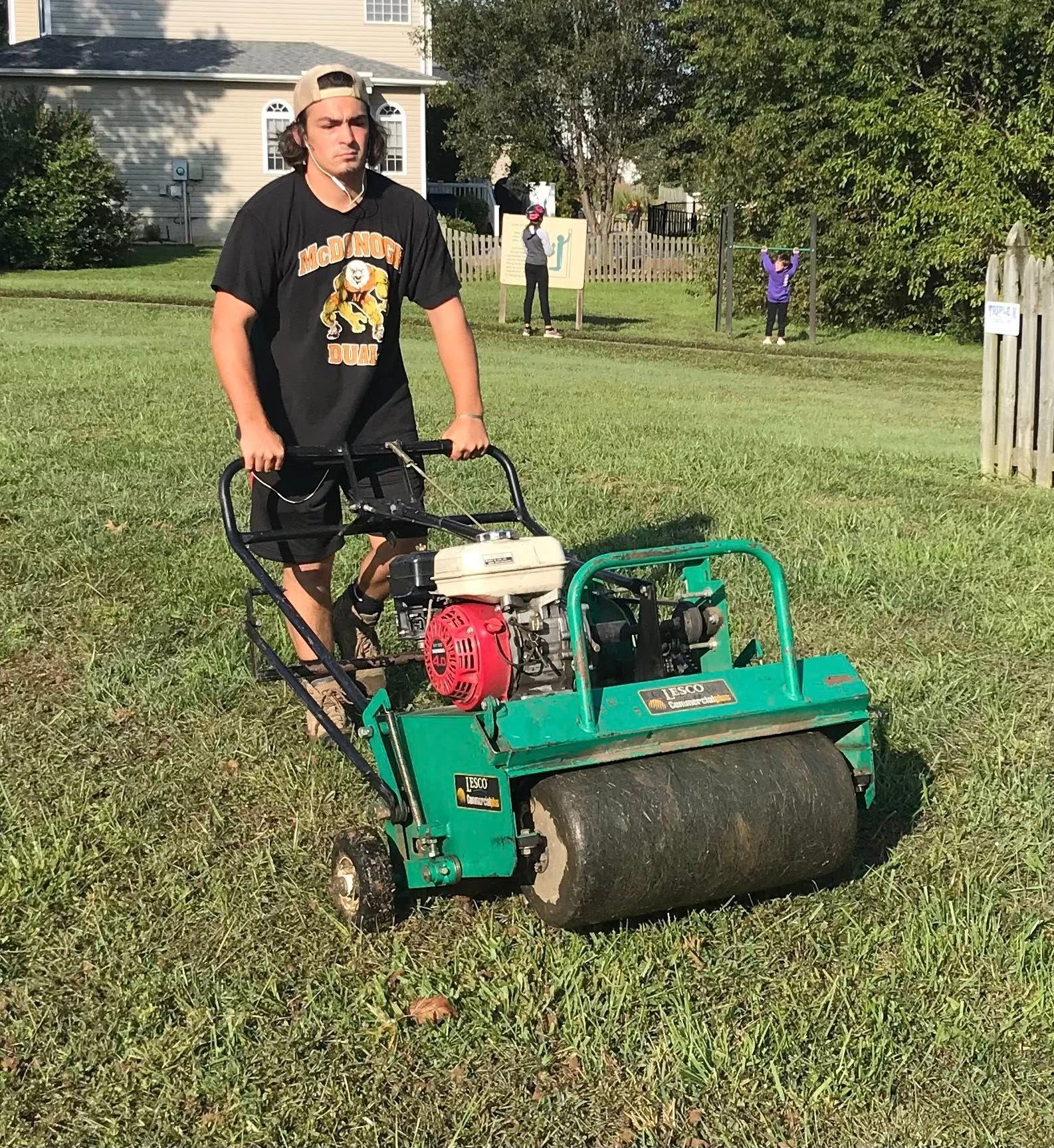 Man standing beside a green lawn roller on a grassy yard, with houses and trees in the background