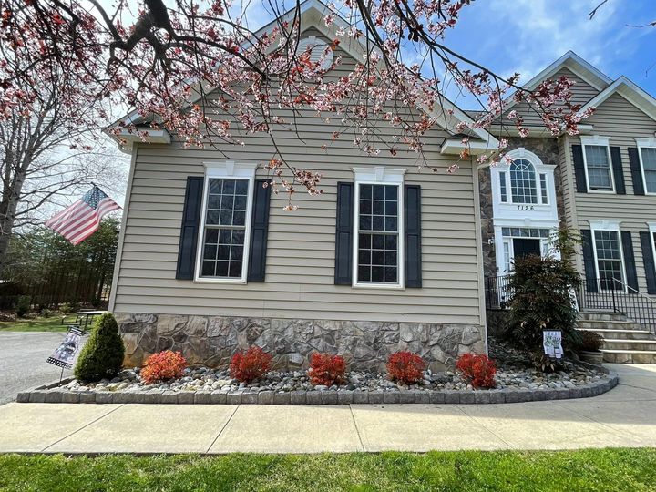 Suburban house with beige siding, black shutters, and blooming pink tree branches overhead.